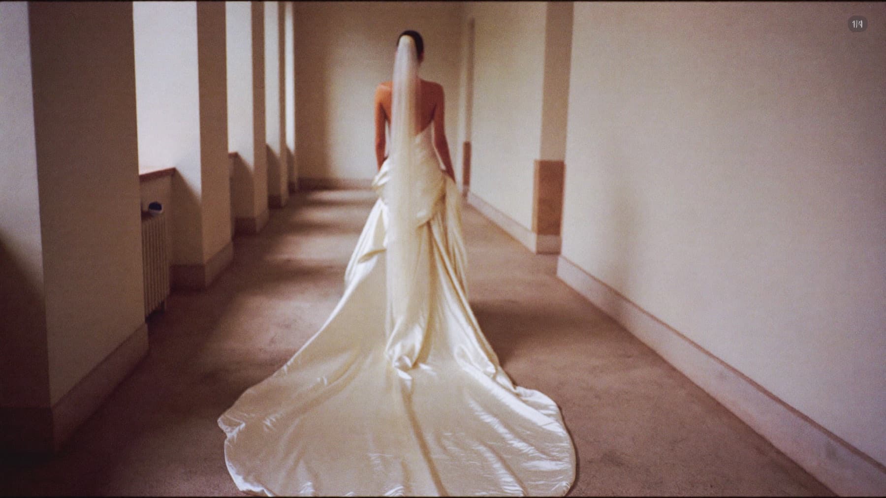 Une mariée de dos s'éloignant dans un long couloir de pierre, sa traîne de soie derrière elle